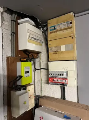 Angle de pièce avant travaux : trois coffrets superposés, dont deux au capot jauni par l’âge, un quatrième sur panneau de bois avec le compteur, gaines et câbles en apparent.