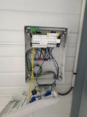 Coffret modulaire ouvert sur un bardage de garage, conducteurs jaunes et bleus raccordés, capot rabattu contre le mur.