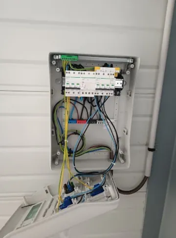 Coffret modulaire ouvert sur un bardage de garage, conducteurs jaunes et bleus raccordés, capot rabattu contre le mur.