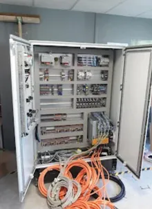 Armoire industrielle sur pied, portes ouvertes, plusieurs rangées d’appareillage et câbles orange encore déroulés au sol.