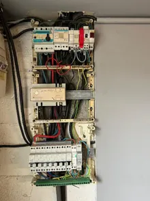Tableau ancien ouvert sur un mur en plâtre : appareillage d’origine en partie haute, protections modulaires ajoutées en partie basse.