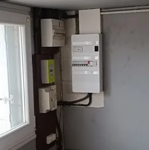 Tableau électrique et compteur installés sur un pan de mur près d’une fenêtre, liaisons apparentes descendant vers le bas du mur.