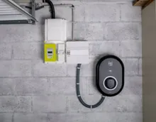 Borne de recharge murale dans un garage : coffret ovale à prise de recharge, relié par une gaine annelée au tableau voisin, à côté du compteur électrique, sur un mur en parpaing brut.